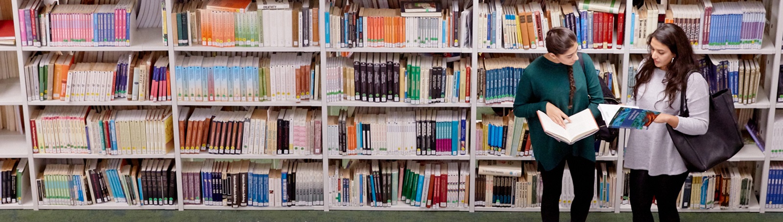 Two students standing in front of a long row of bookshelves discussing a book they are holding